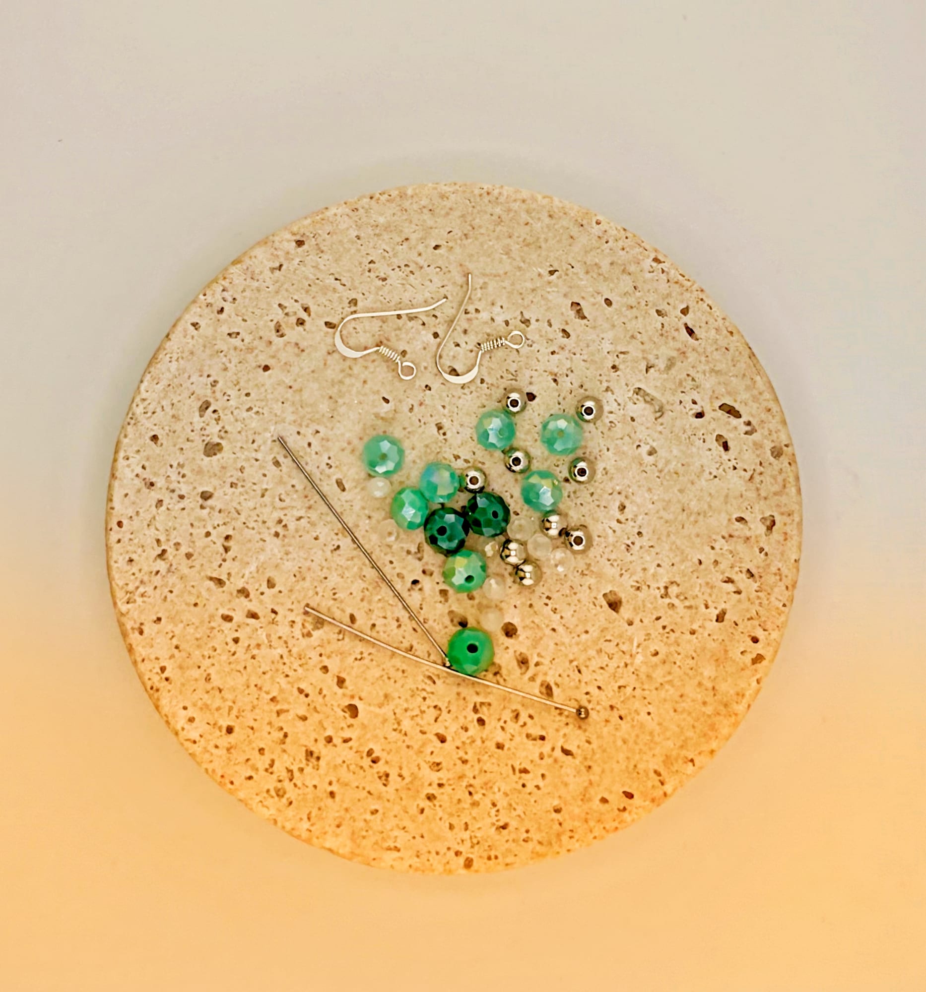 Vue de dessus d'un rond de liège : deux crochets d'oreille argentés, un petit tas de perles vertes facettées et deux tiges métalliques croisées